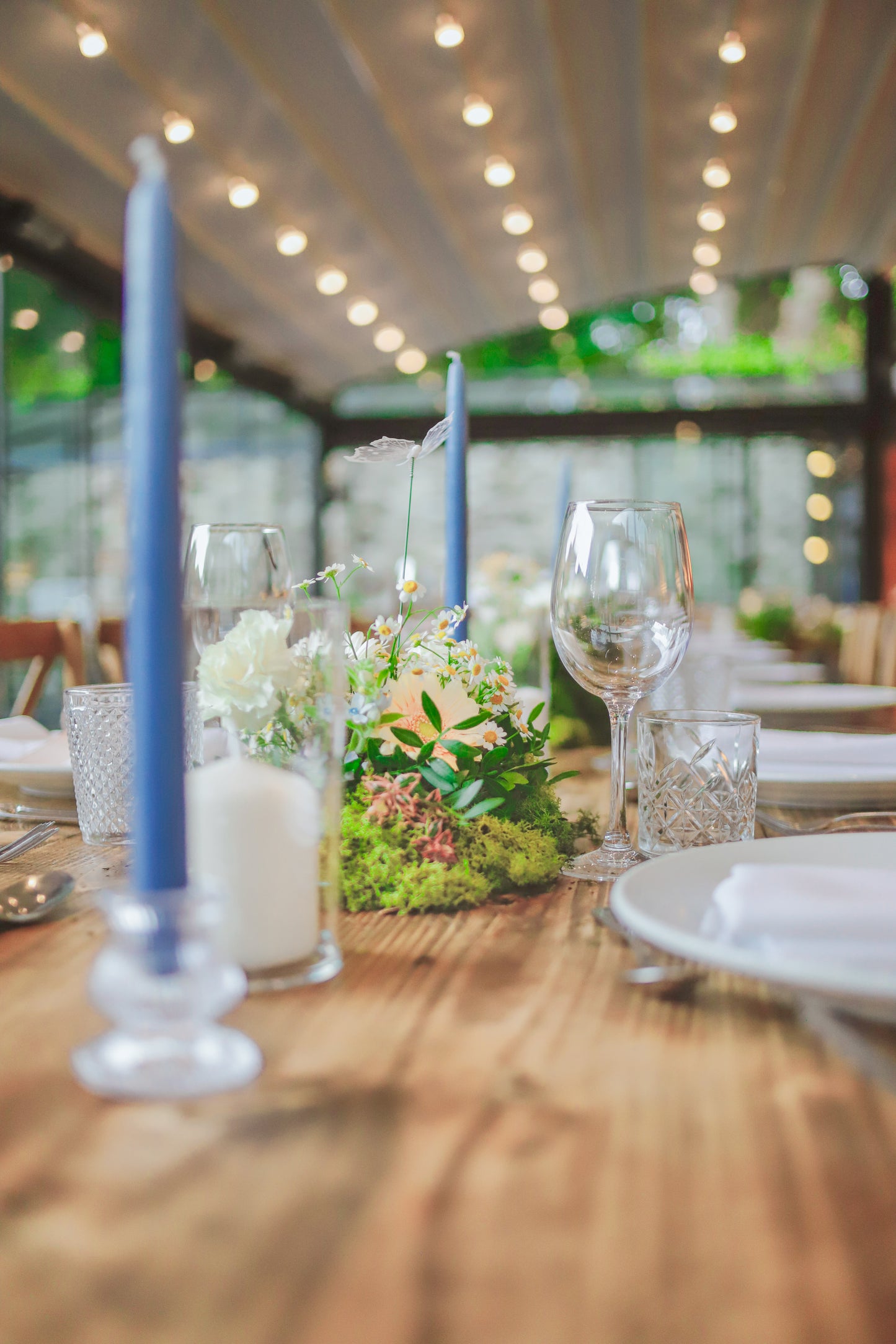 Table setting featuring a small moss floral arrangement, blue taper candles, ivory pillar candles and glasses.