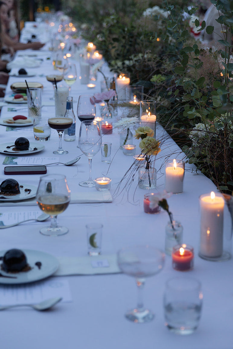 An outdoor evening wedding reception table setting with guests seated on both sides, a long white tablecloth, lit candles, wine glasses, plates, and floral centerpieces.