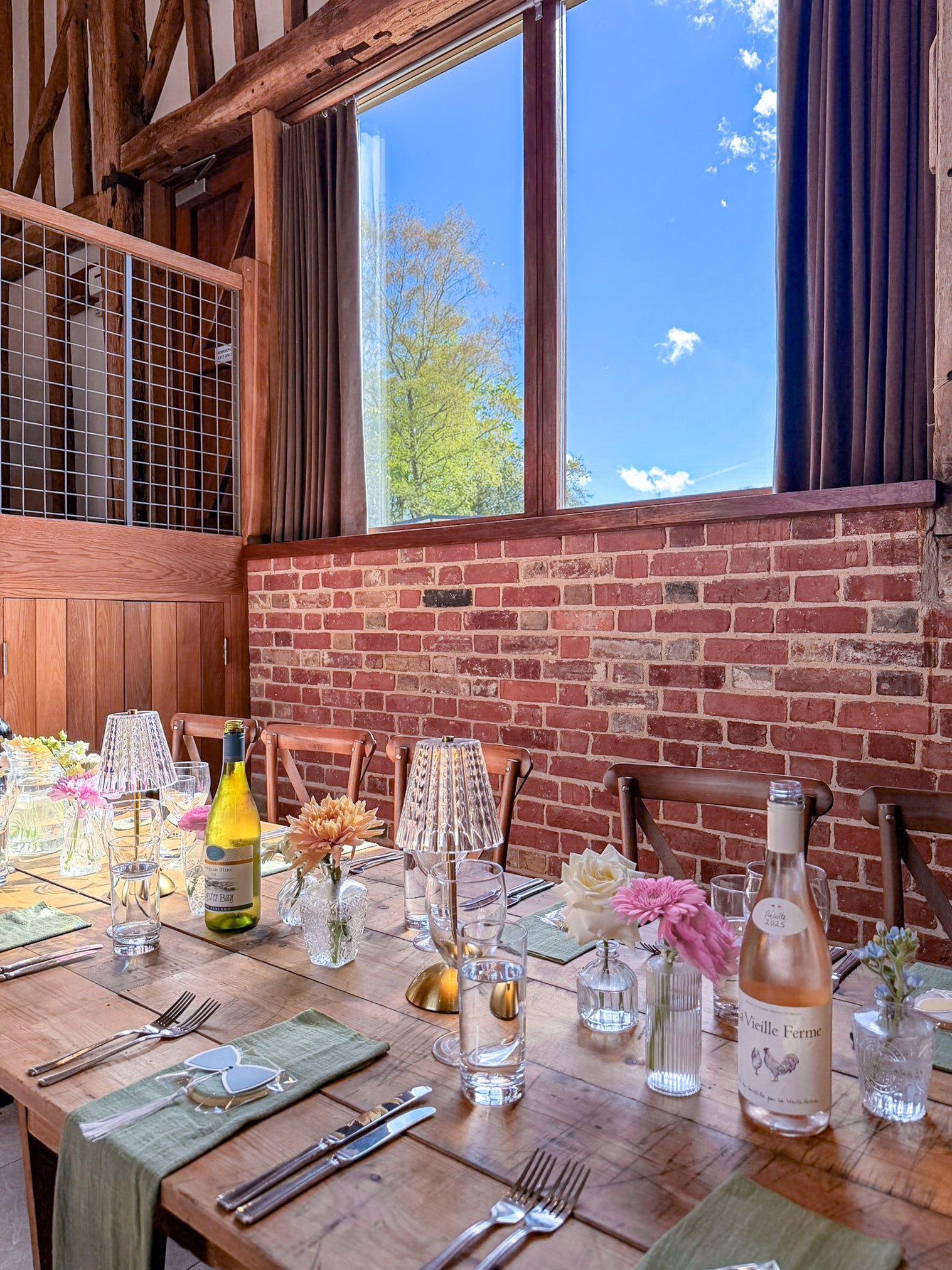 Dining table set for a meal with wine bottles, glasses, and flowers in a rustic setting with a brick wall and large window.