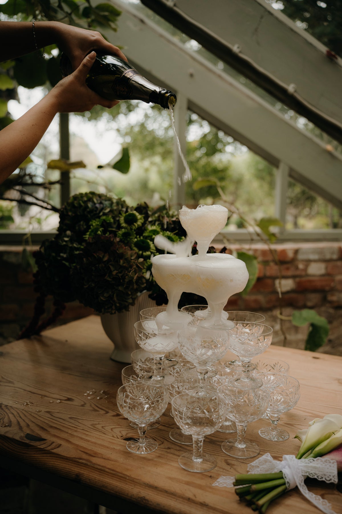 A champagne tower made of stacked clear champagne glasses on a wooden table, with a bottle being poured into the center. A person's arm and hand are visible pouring the liquid, with a greenhouse or garden interior in the background.