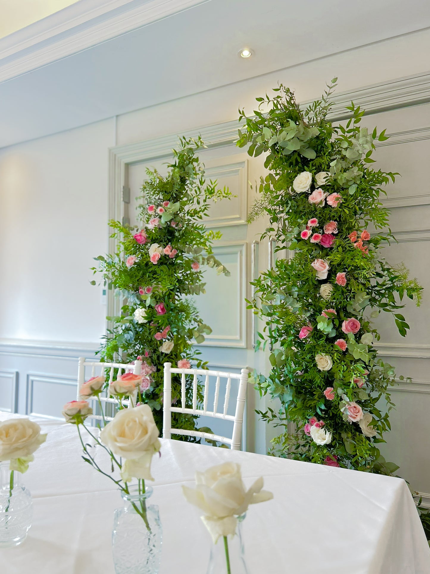 Floral arches with pink and white flowers in a room with white walls and chairs.