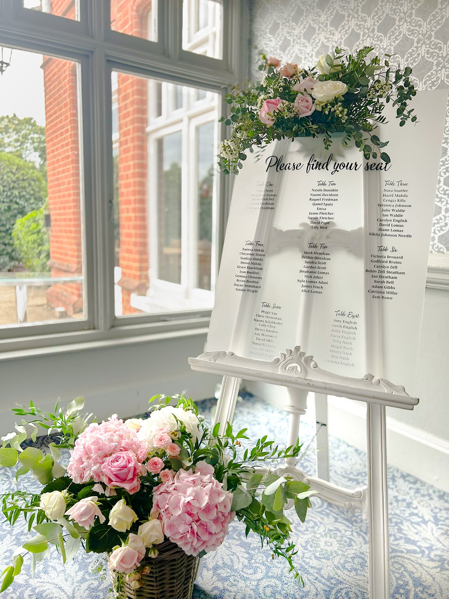 Decorative setup with floral arrangements and a seating chart in a room with large windows.