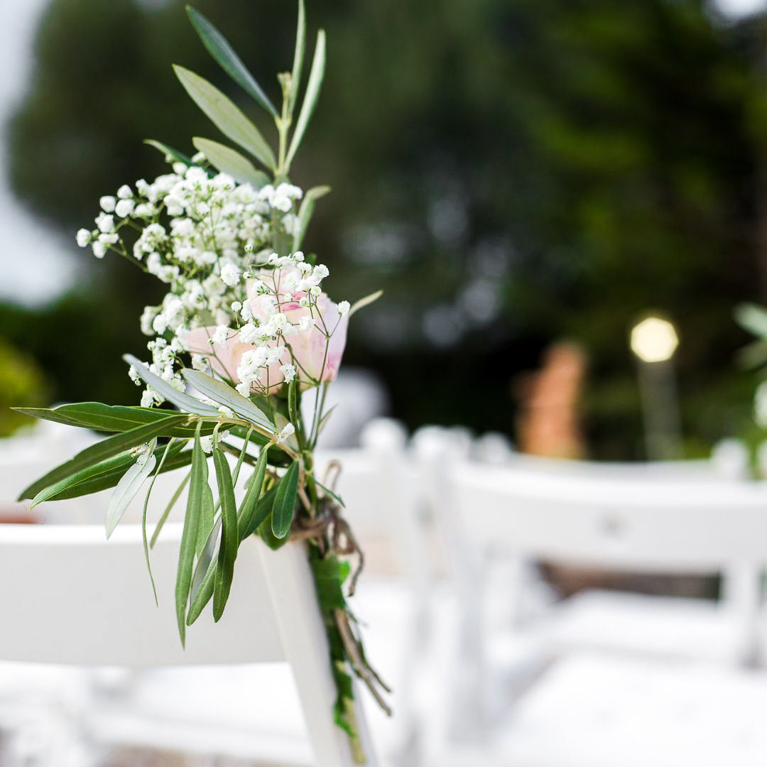 Floral arrangement with white flowers and green leaves on a white chair at a wedding outdoor. 