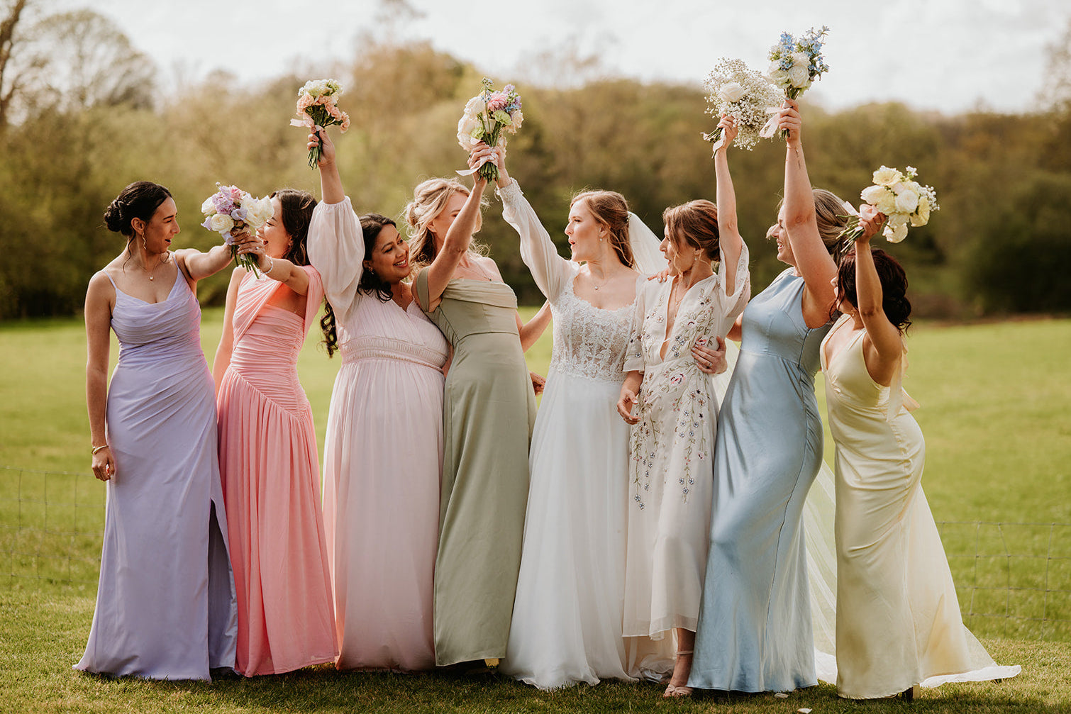 Group of bridemaids and bride in colorful dresses standing in a field, holding flowers.